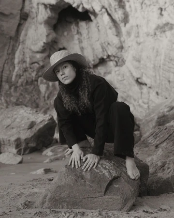Monochrome portrait of a young woman with curly hair wearing a straw hat and dark clothing, crouching on a rock with a rock cave behind.