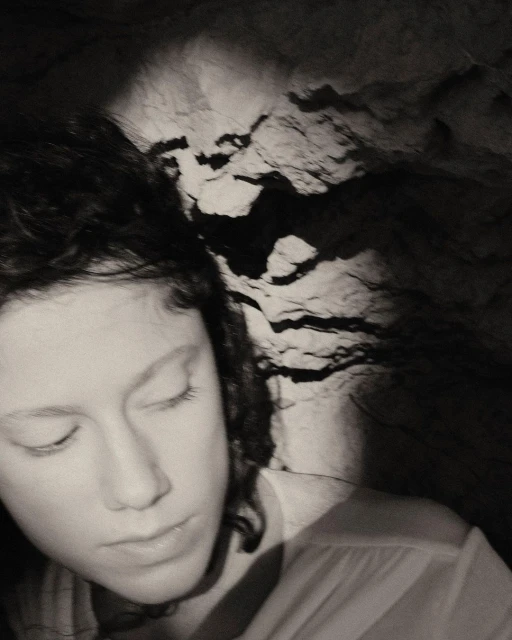 Monochrome portrait of a young woman with closed eyes, set against a textured rock background in a dimly lit, atmospheric scene.