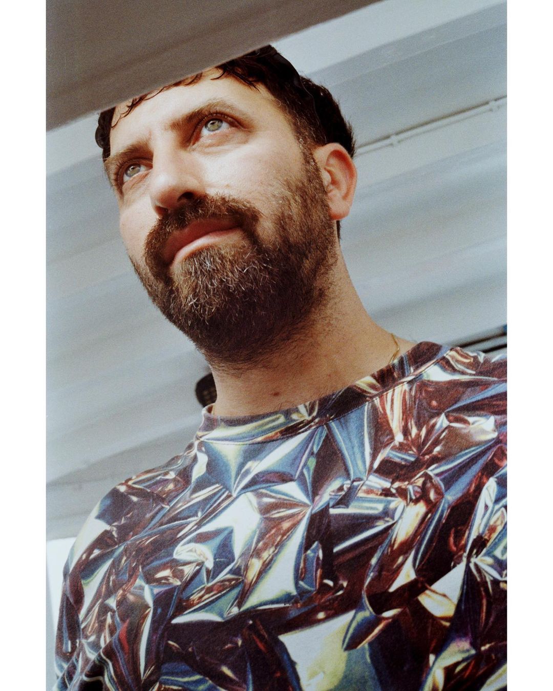 Low-angle portrait of a man with a beard wearing a metallic foil-patterned shirt against a white ceiling background.