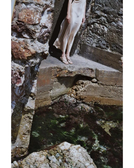 An adult female in a dress stands on a concrete structure beside the ocean. Light reflects in the water. A rocky wall borders the scene.