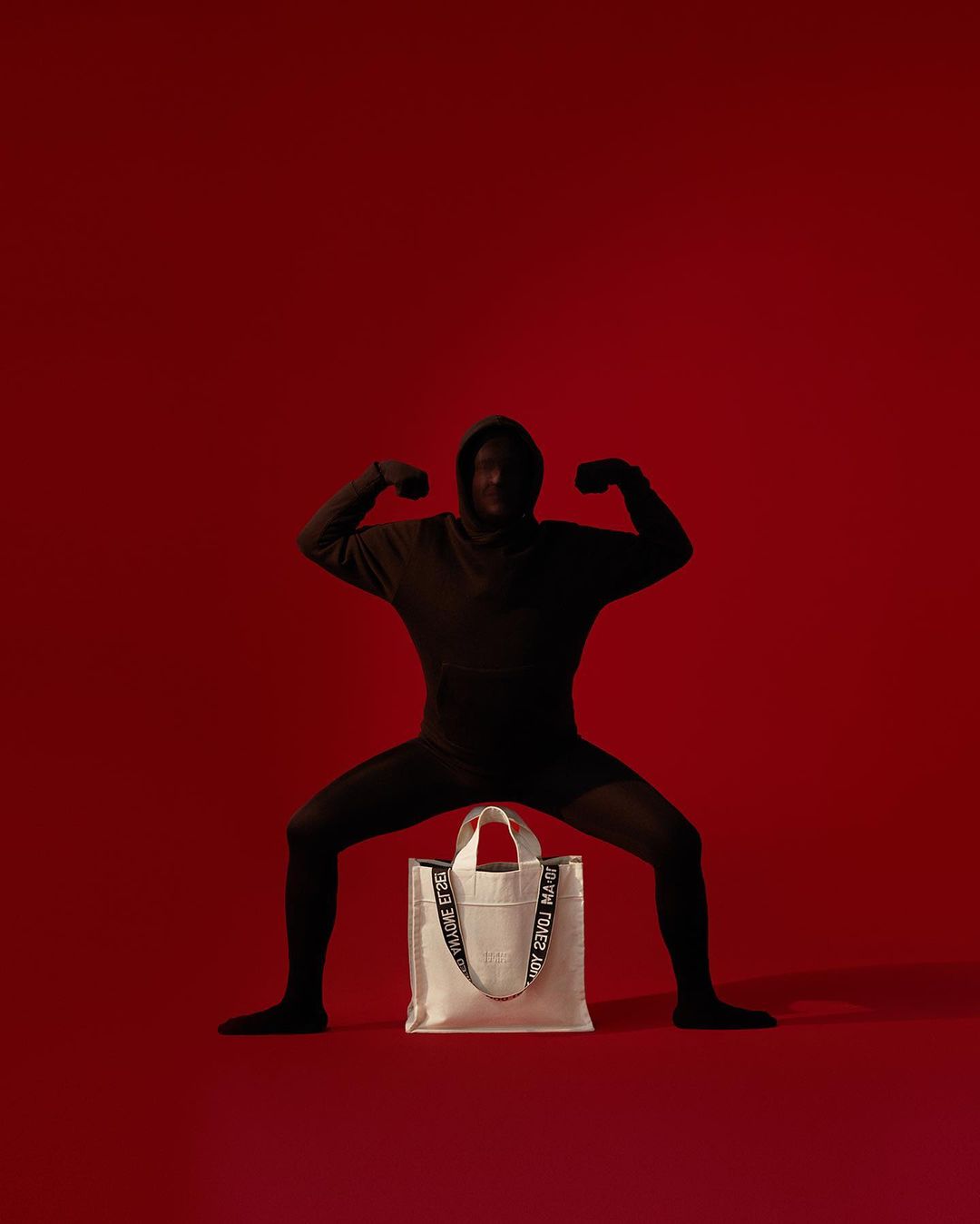 A person in a black hooded outfit crouching over a white tote bag against a solid red studio background.