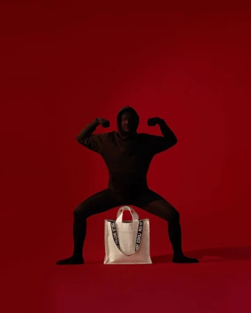 A person in a black hooded outfit crouching over a white tote bag against a solid red studio background.