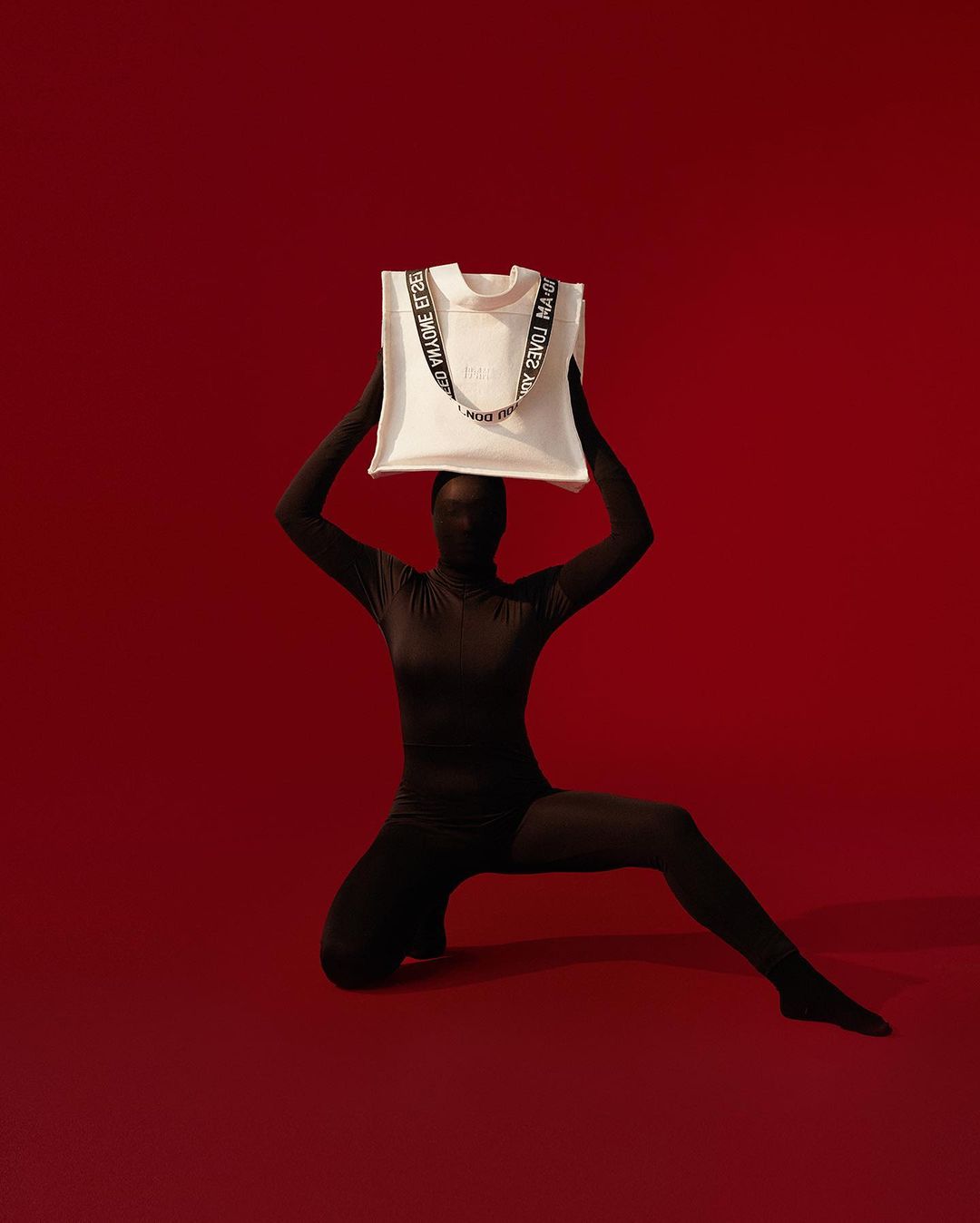 Person in dark bodysuit poses in front of red background holding a cream-colored tote bag with black text strap above their head.