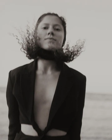 Monochrome portrait of a person with curly hair blowing in the wind wearing a dark open blazer against a bright sky.