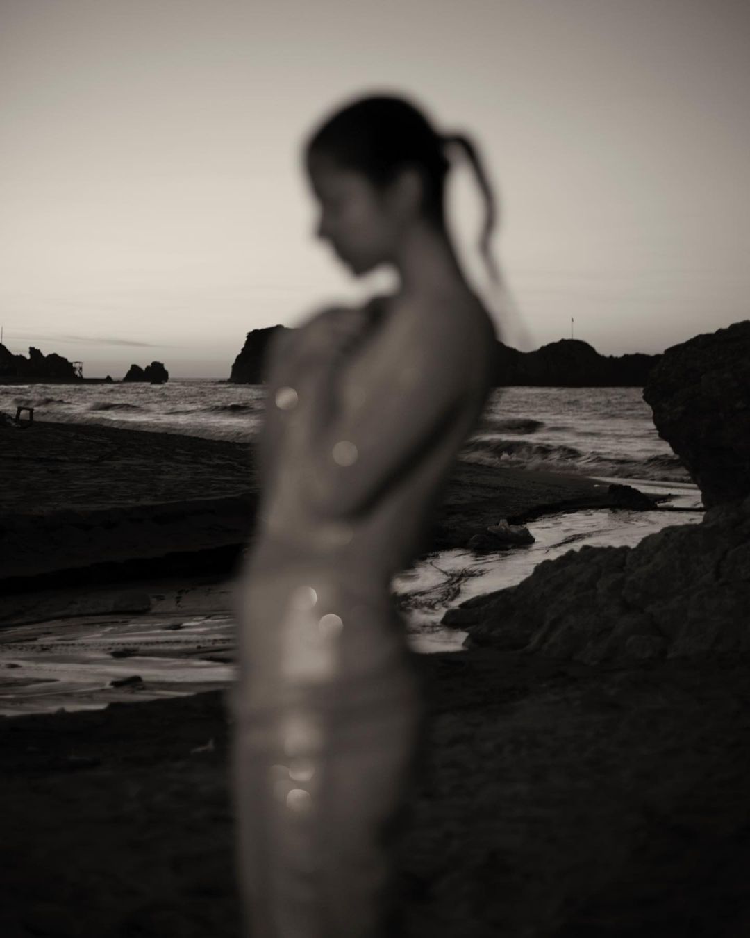 Out of focus portrait of a woman standing on a sandy beach near the ocean at sunset with rocky outcrops in the background.