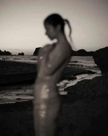 Out of focus portrait of a woman standing on a sandy beach near the ocean at sunset with rocky outcrops in the background.