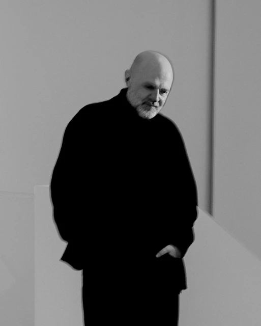 Portrait of a mature adult man with a beard wearing a black sweater, standing indoors against a neutral grey background, with natural lighting.