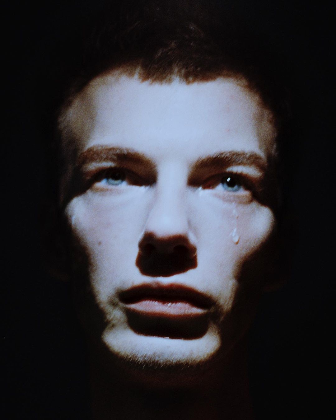 Close-up portrait of a young adult male with a tear rolling down his cheek, set against a dark, contrasting background with dramatic lighting.