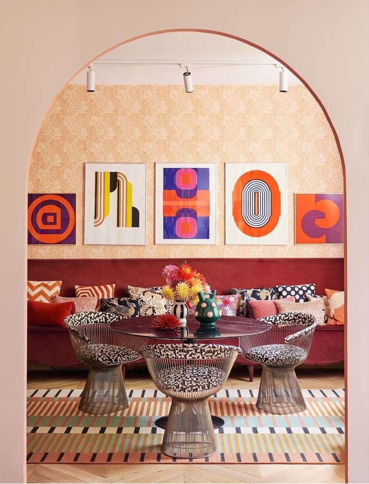 Eclectic dining area with round table, patterned chairs, colorful geometric art, and decorative cushions against a peach-toned backdrop.
