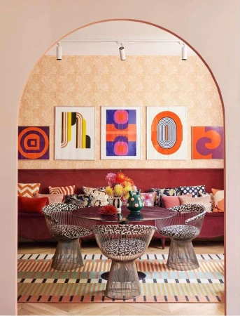 Eclectic dining area with round table, patterned chairs, colorful geometric art, and decorative cushions against a peach-toned backdrop.
