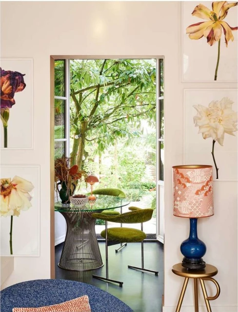 Living room corner with a modern glass table, green chairs, flower pictures, and a lamp on a table, creating a stylish home interior.