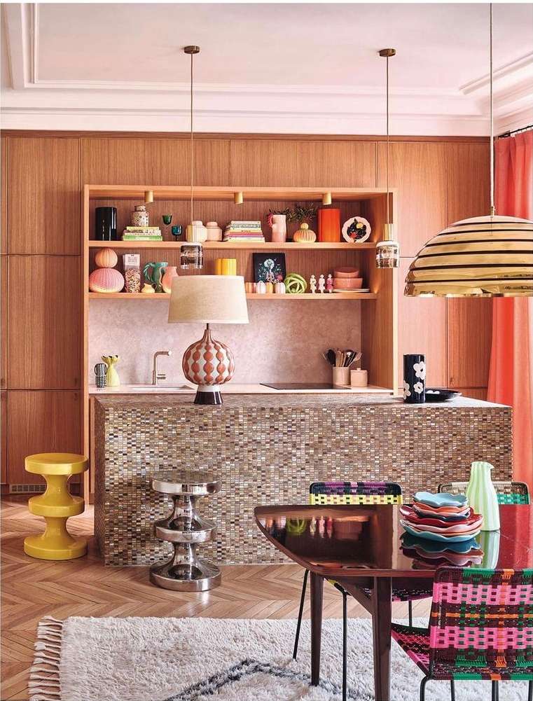 Bright kitchen interior with wood cabinets, mosaic island, pendant lighting, colorful chairs, and a patterned rug on a herringbone floor.
