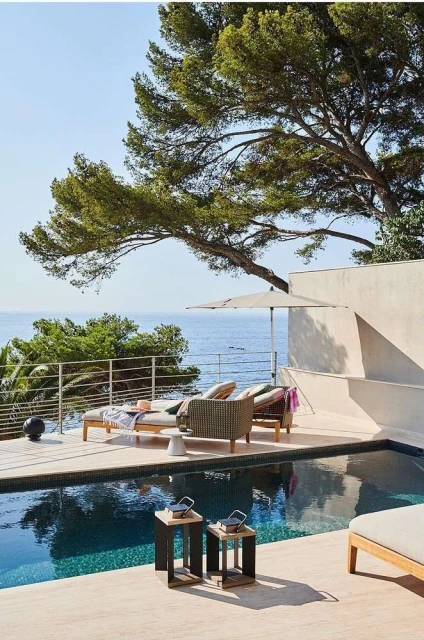 Outdoor poolside patio featuring lounge chairs, side tables, and a large tree providing shade in a tranquil setting.