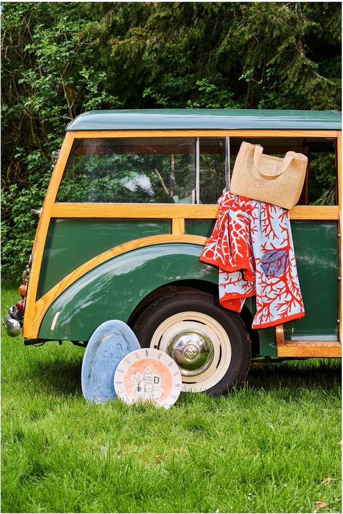 Vintage green woody car with straw bag and colorful towel hanging, parked on green grass with vintage plates in an outdoor setting.