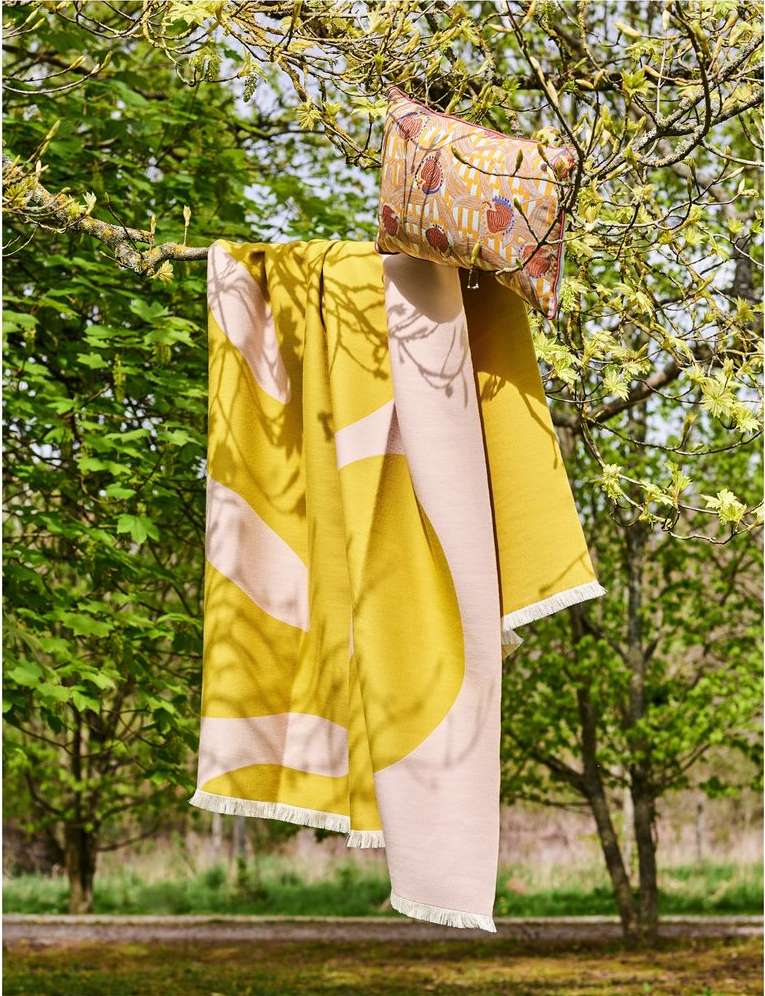 Yellow and pink blanket with fringe and patterned pillow hanging from tree branch outdoors with green trees in the background.