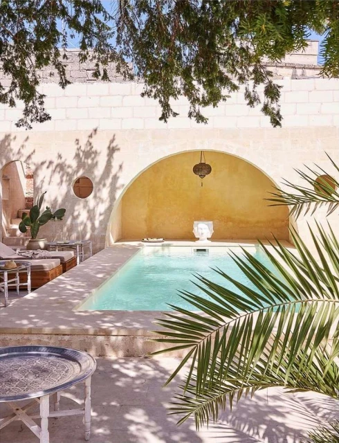 Outdoor pool area with light turquoise water, a stone deck, palm fronds in foreground, an arched alcove, and dappled sunlight.
