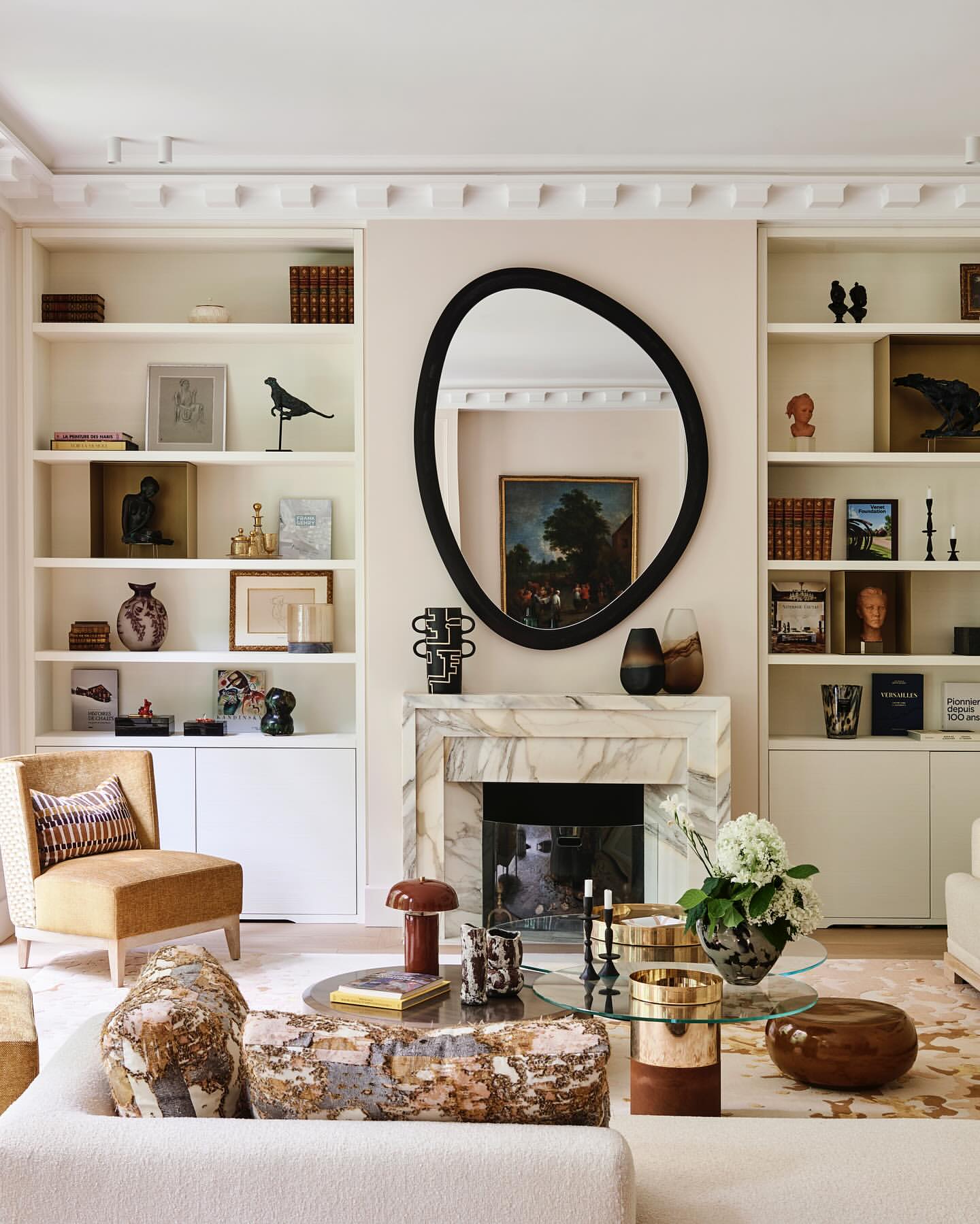 Interior design of a living room featuring neutral tones, bookshelves flanking a marble fireplace, and a sculptural mirror above the mantle.