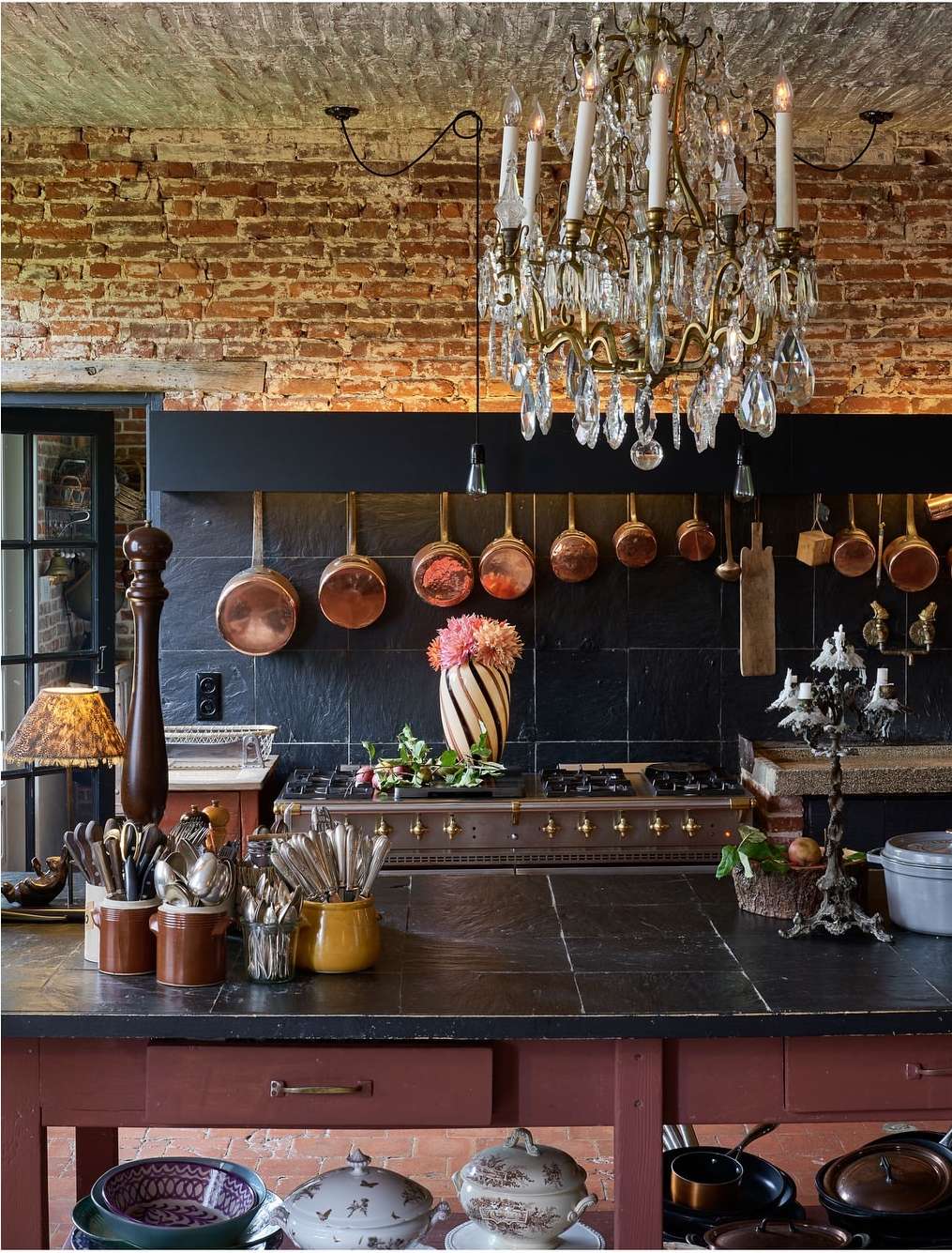 Chandelier hangs above a kitchen counter with copper pots, silverware, flower-filled vase, and a red brick wall at the back.