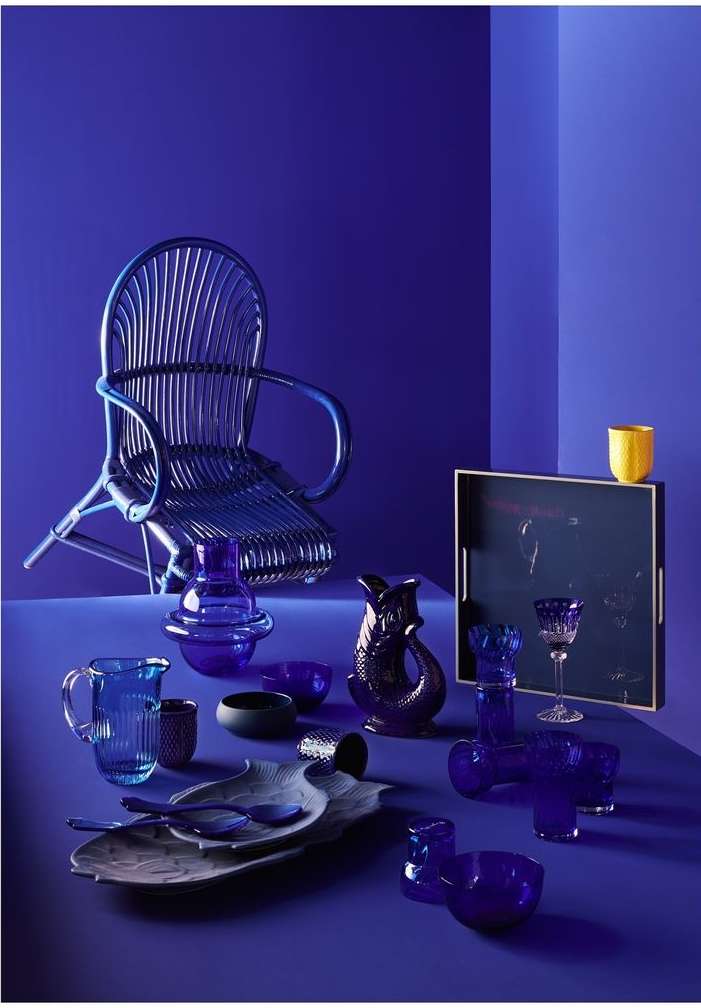 Still life studio shot of assorted blue glassware, tableware, and a rattan chair against a monochromatic royal blue backdrop.