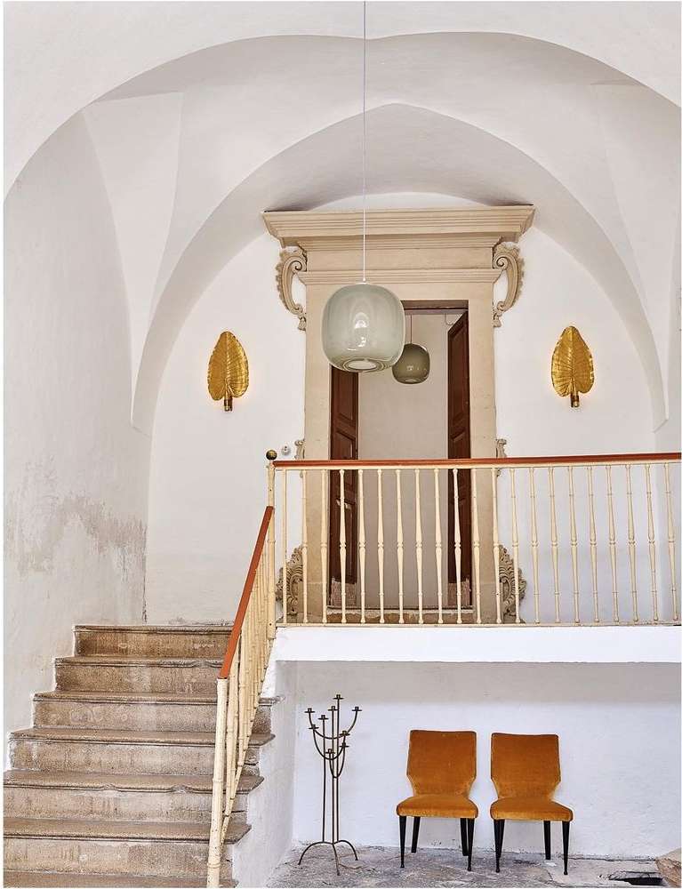 Interior view of a white building featuring a staircase, two chairs, and architectural lighting fixtures in a minimalist aesthetic.