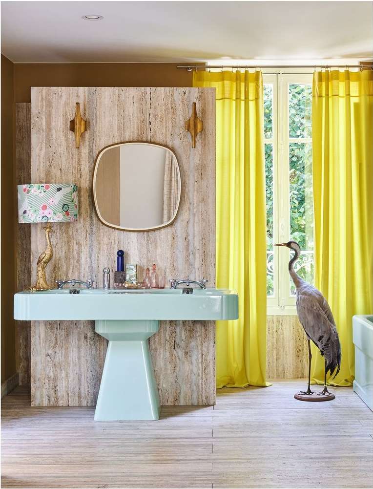 Bathroom interior with light blue double sink, gold accents, travertine wall, yellow curtains, window view and heron statue.