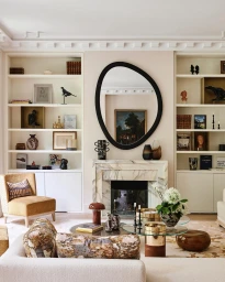 Interior living room featuring a marble fireplace, asymmetrical black-framed mirror, built-in bookshelves, and modern furniture.