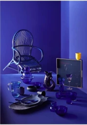 Monochromatic still life featuring assorted blue glassware, plates, and a matching rattan chair against a deep blue wall.