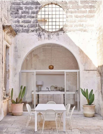 Outdoor dining area with white table and chairs in stone courtyard with potted cactus plants against a rustic arched wall