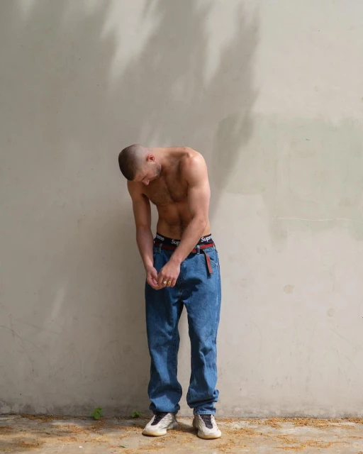 A shirtless adult male wearing jeans and white sneakers stands against a pale wall in natural light, with tree shadows above him.