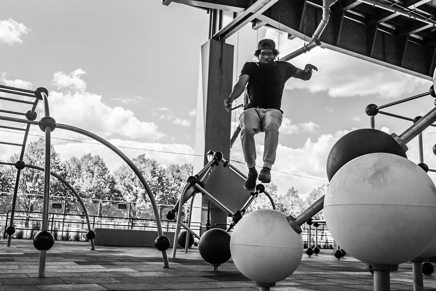 Adult male jumping between steel playground structures in an outdoor black and white monochrome photograph with a cloudy sky.