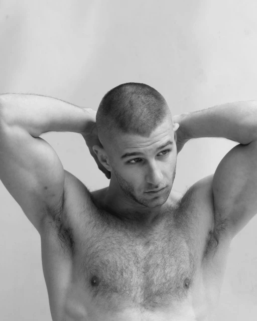 Black and white studio portrait of a muscular adult male with short hair, shirtless, posing with his hands behind his head.