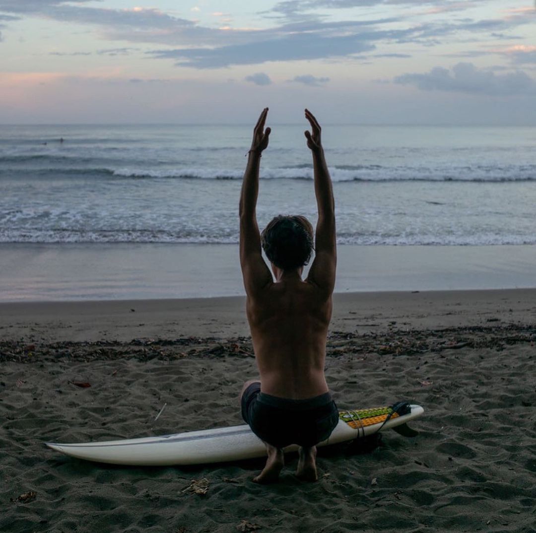 Person in a squatting yoga pose on a sandy beach next to a surfboard at dusk with the ocean and cloudy sky in the background.
