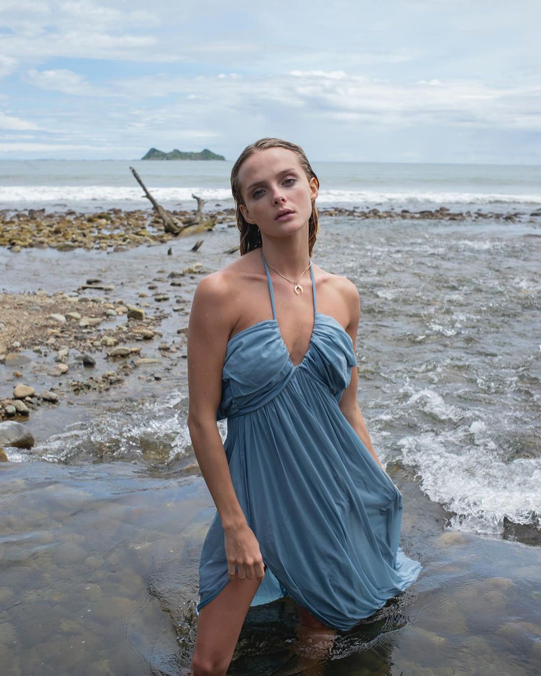 Adult female in a halter dress standing in shallow water at a rocky beach with a small island in the background under a cloudy sky.