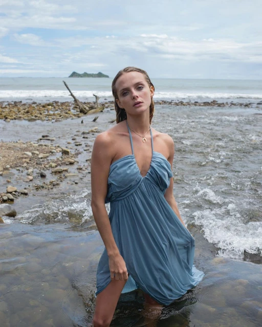 Adult female in a halter dress standing in shallow water at a rocky beach with a small island in the background under a cloudy sky.