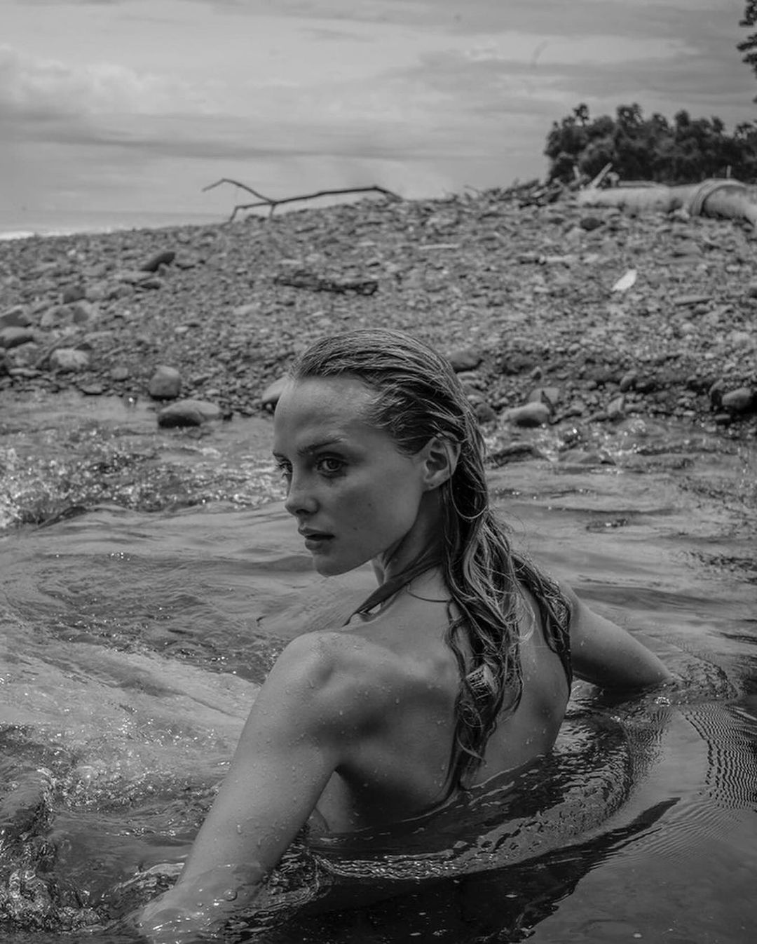 Black and white image of a blond woman in water looking over her shoulder on a rocky beach with a cloudy sky.