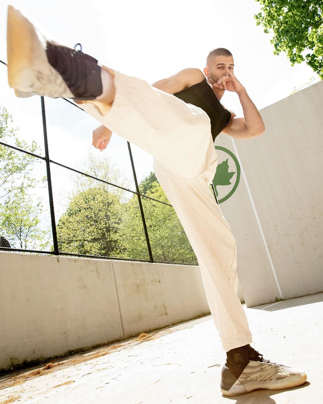 Athletic man in white pants and black tank top stretches his leg high outdoors with greenery and a building in the background on a sunny day.