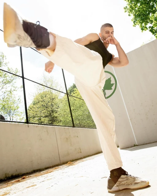 Athletic man in white pants and black tank top stretches his leg high outdoors with greenery and a building in the background on a sunny day.