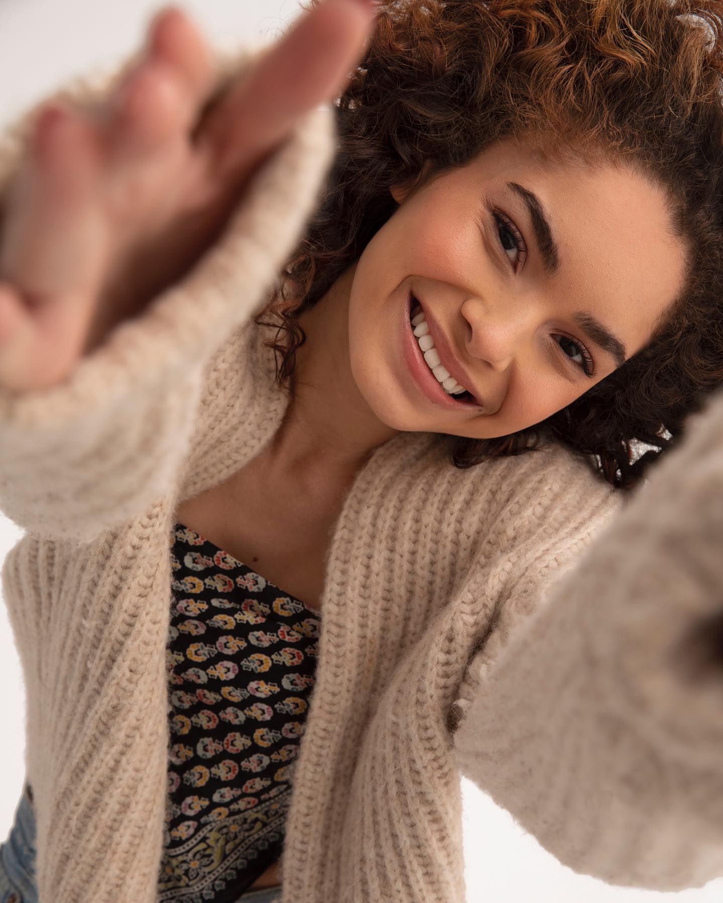A woman with curly hair smiles and reaches toward the viewer, wearing a beige cardigan and a patterned top in a bright indoor space.