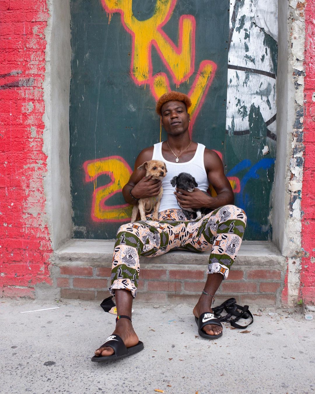Man sitting with two dogs in front of graffiti covered wall. He is wearing a white tank top, patterned pants, and black sandals.
