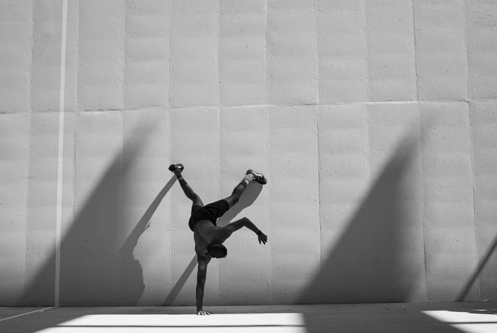 Monochrome image of adult male doing a one-handed handstand against a concrete wall with shadows, shot in an outdoor setting.