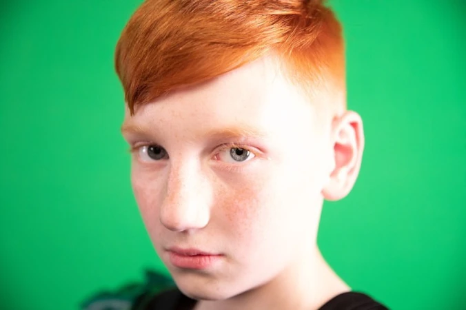 Close-up portrait of a young person with bright red hair and freckles against a vibrant green background.