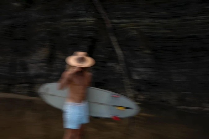 Blurred motion shot of a shirtless adult male carrying a surfboard and wearing a hat, near a dark rocky outcropping.