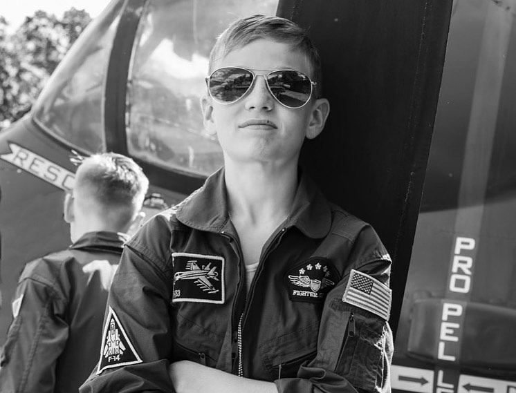 A black and white shot captures a confident young boy in aviator sunglasses and a fighter pilot jumpsuit posing near an aircraft.
