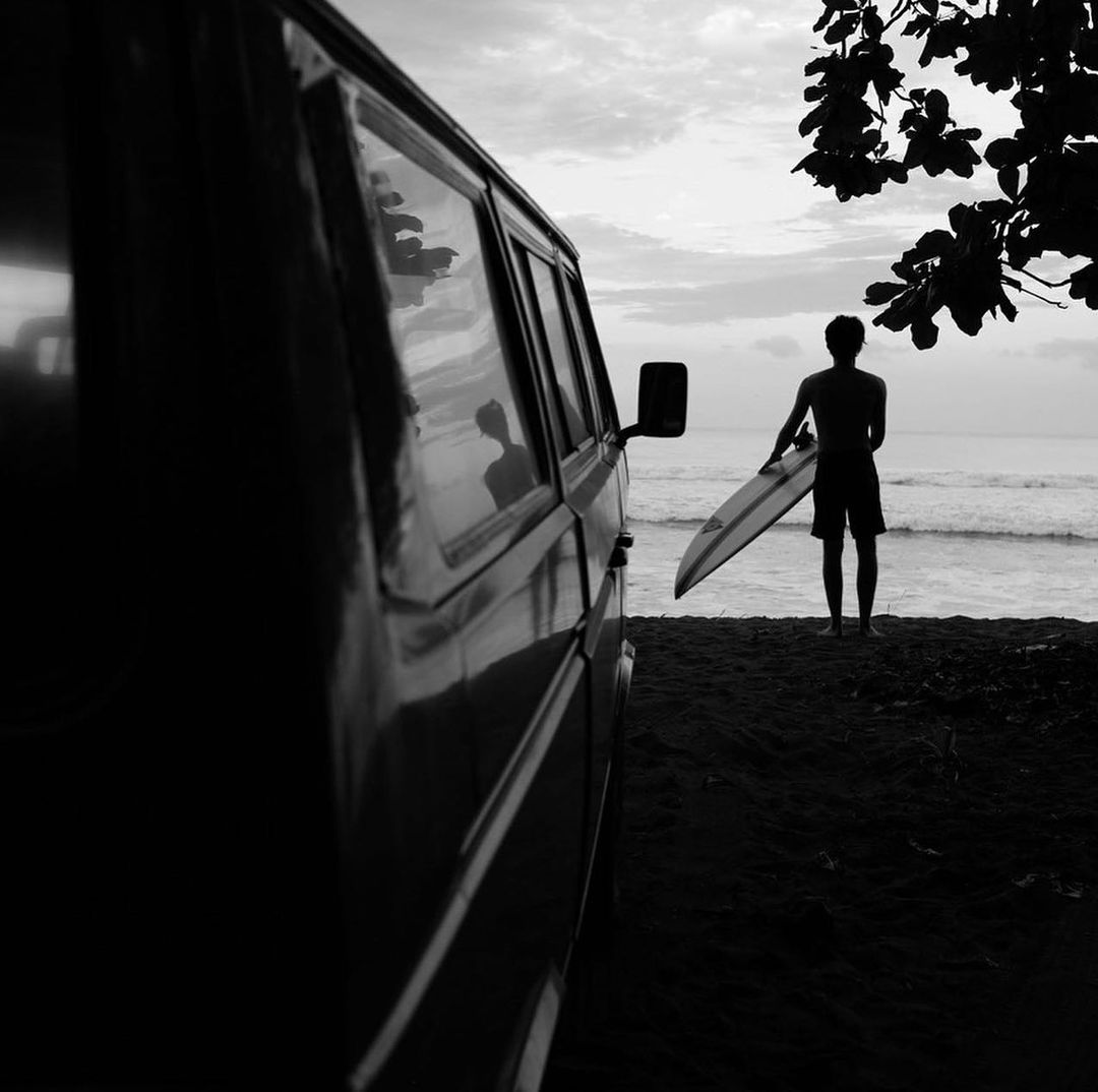 Black and white photo of a man with a surfboard standing near a van on a beach with ocean waves under an overcast sky.