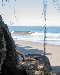 Woman in brown bikini lying on a rocky outcrop on a sandy beach by the ocean, photographed by Didier Gault.