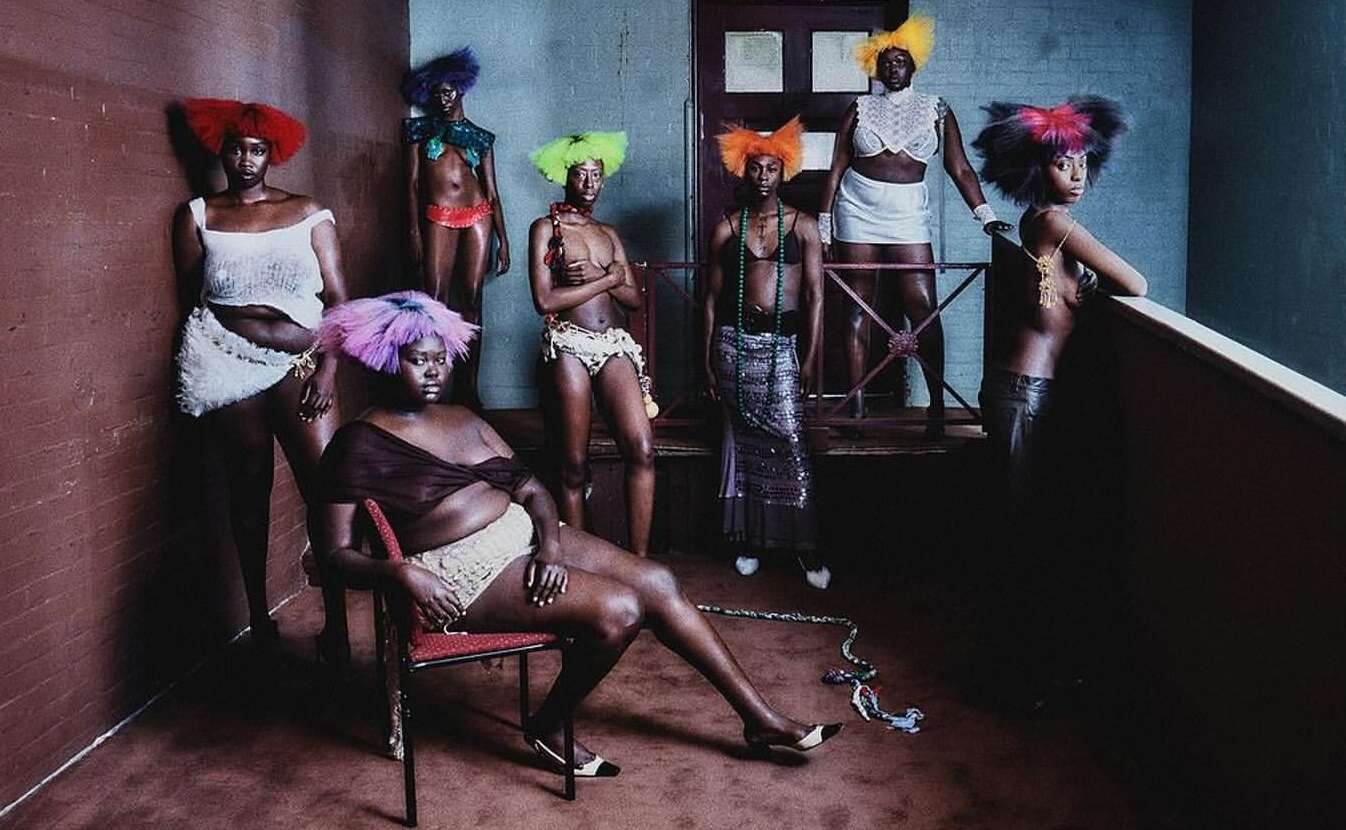 Group of seven African American people in minimal clothing and colorful wigs pose inside a room with brick walls and a balcony.