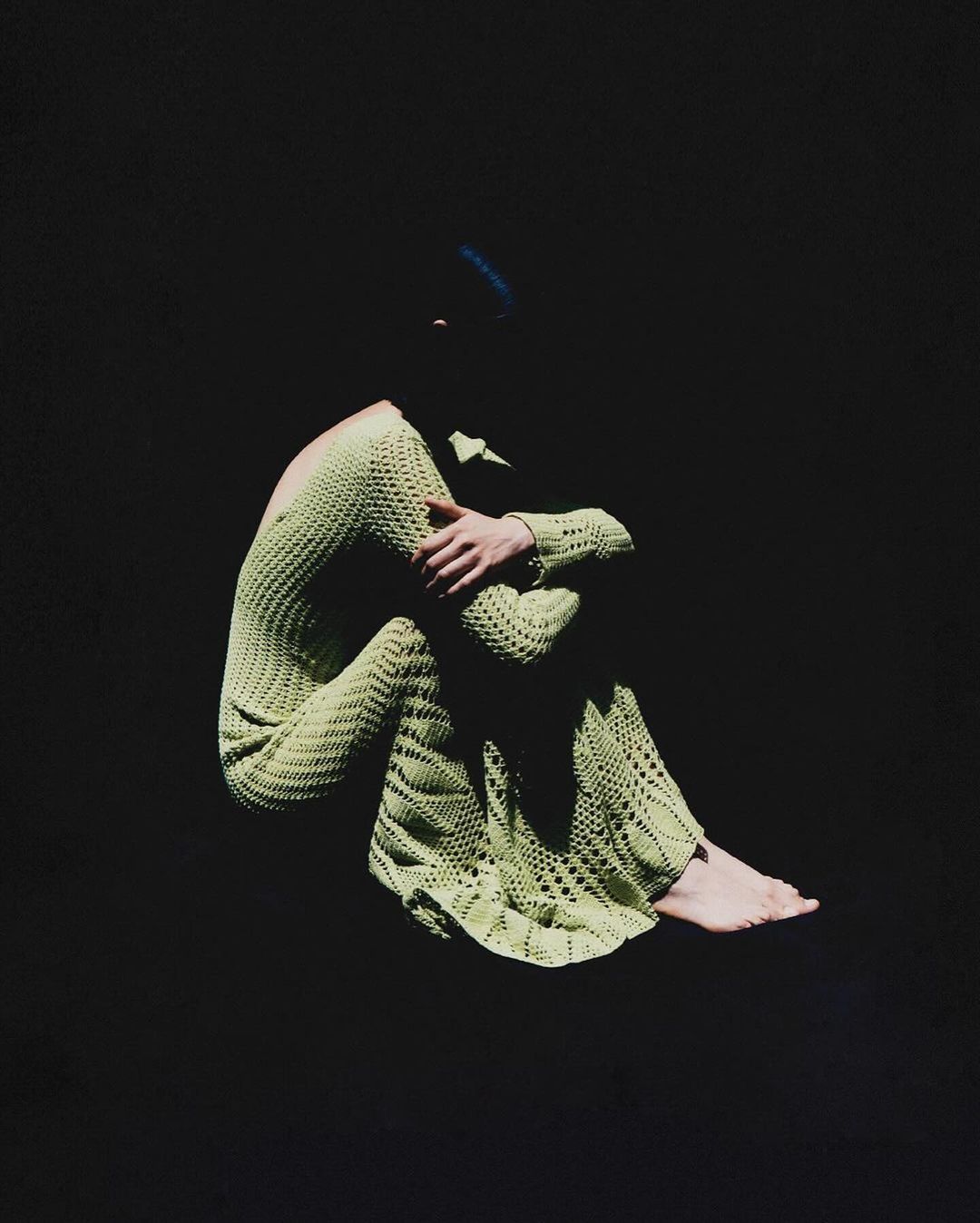 A woman in a green knitted dress sits with arms around her legs against a dark background, creating a portrait with simplicity and style.