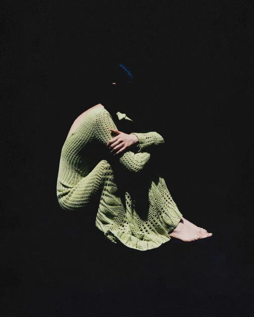 A woman in a green knitted dress sits with arms around her legs against a dark background, creating a portrait with simplicity and style.