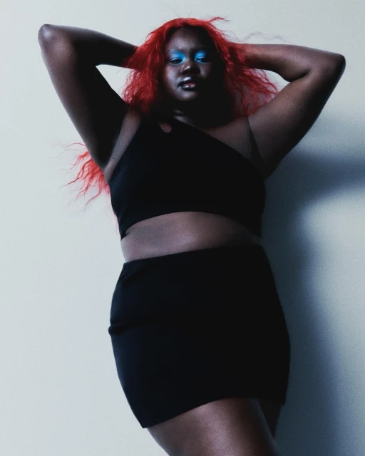 Attractive plus-size woman with red hair and blue eye shadow poses in a black crop top and skirt against a light gray background.