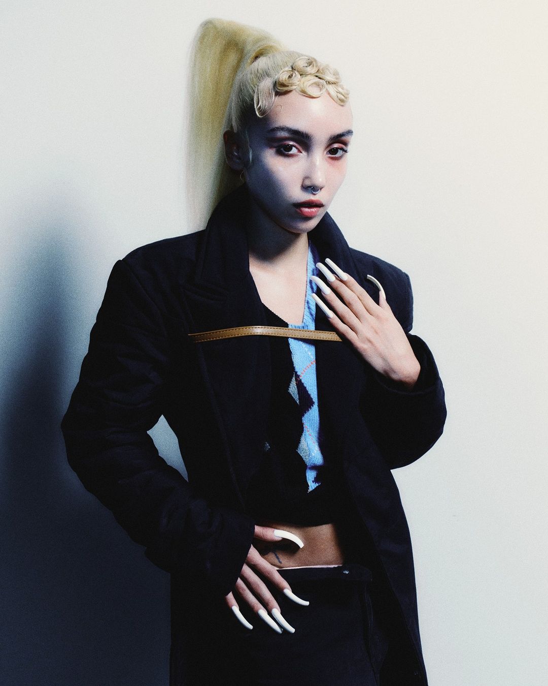 Adult female posing in a dark coat, blue top, and black skirt in front of a neutral background with a high blond ponytail hairstyle.